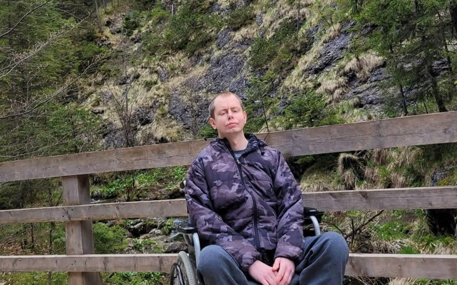 
        Mężczyzna na wózku inwalidzkim odpoczywa na drewnianym moście w otoczeniu natury.
    