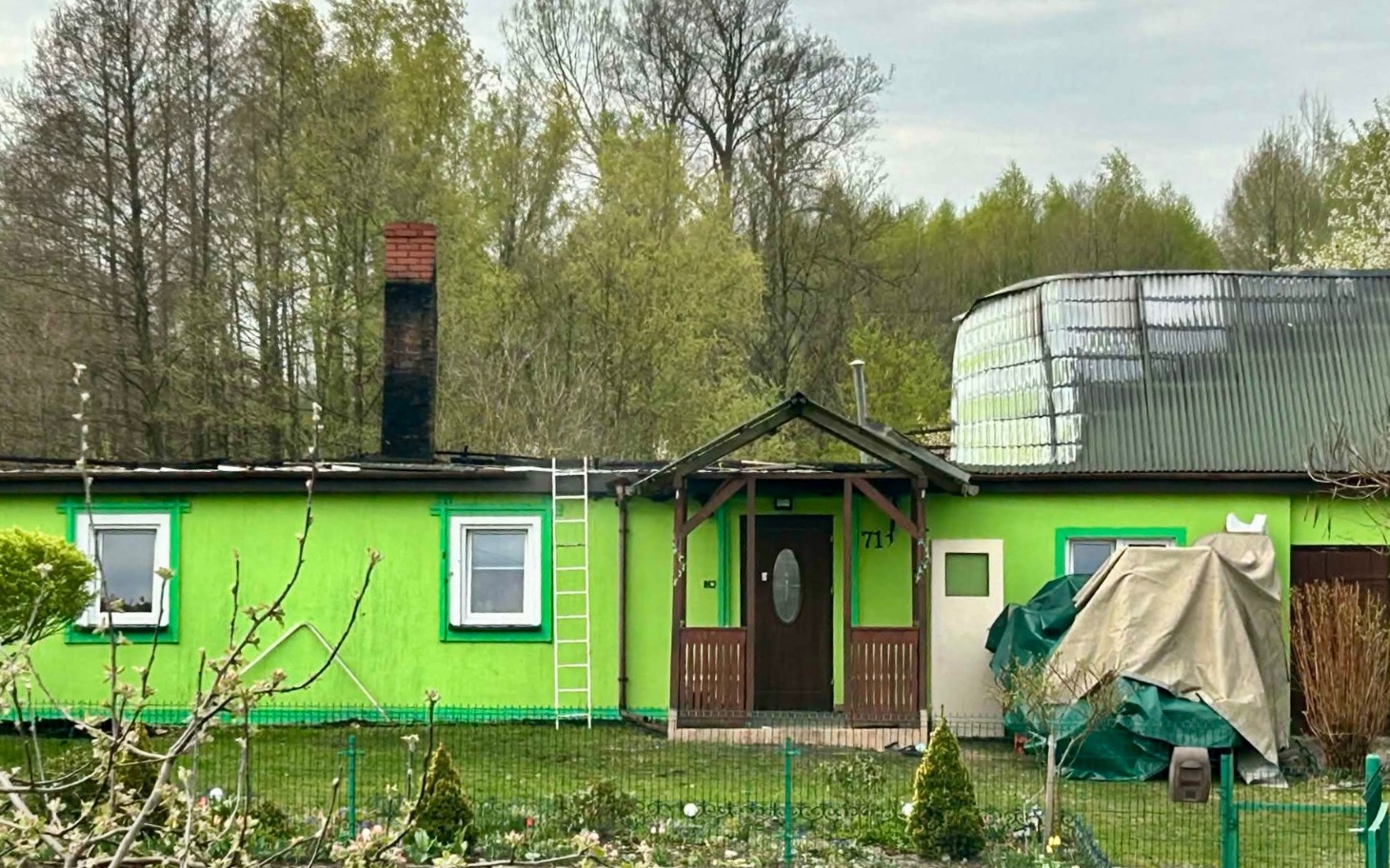 
        Zielony domek z kominem i tarasem w otoczeniu drzew.
    