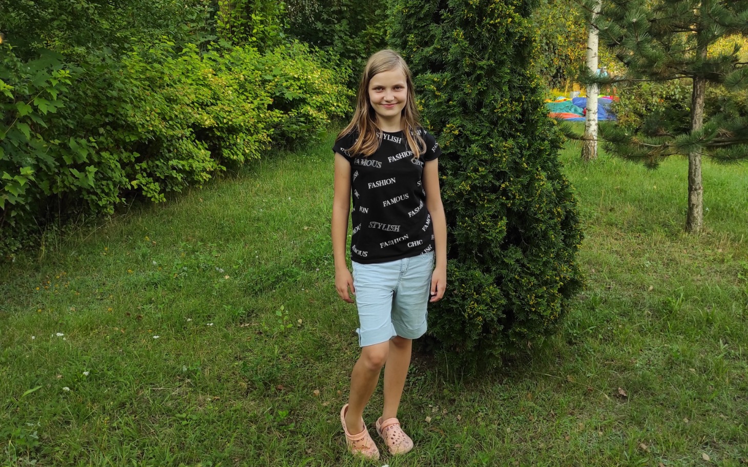 
        Dziewczynka w letnim stroju stoi w ogrodzie, otoczona zielenią.
    