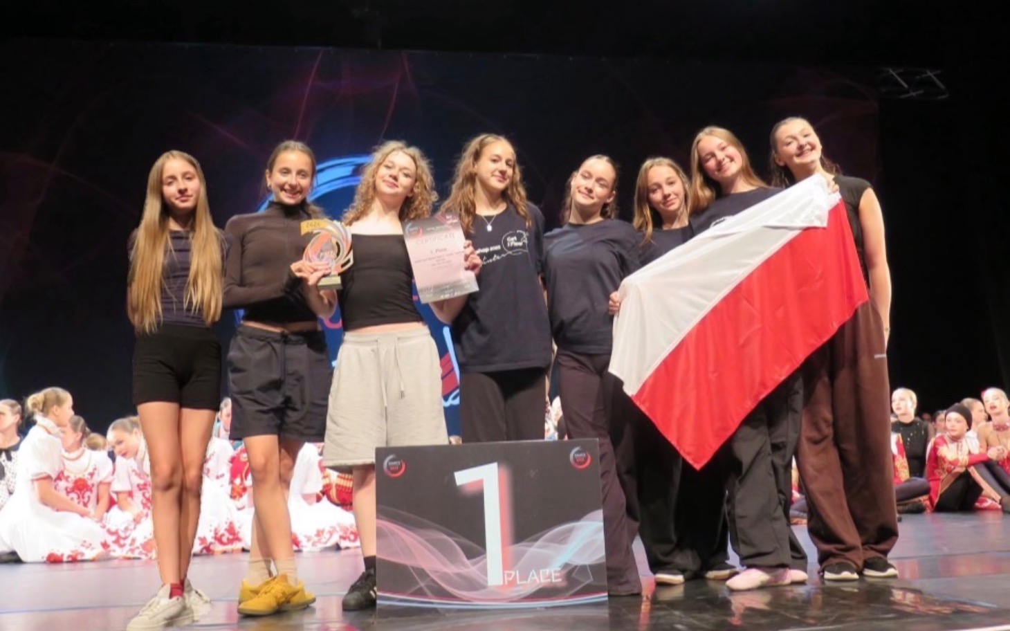 
        Grupa dziewcząt na scenie z nagrodą, trzymających flagę Polski.
    