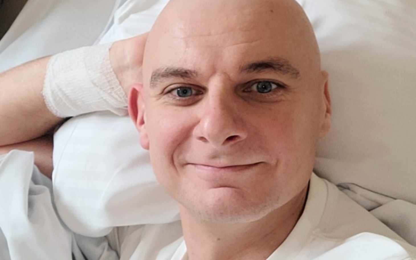 
        Mężczyzna z uśmiechem leży w łóżku, z bandażem na ręce.
    