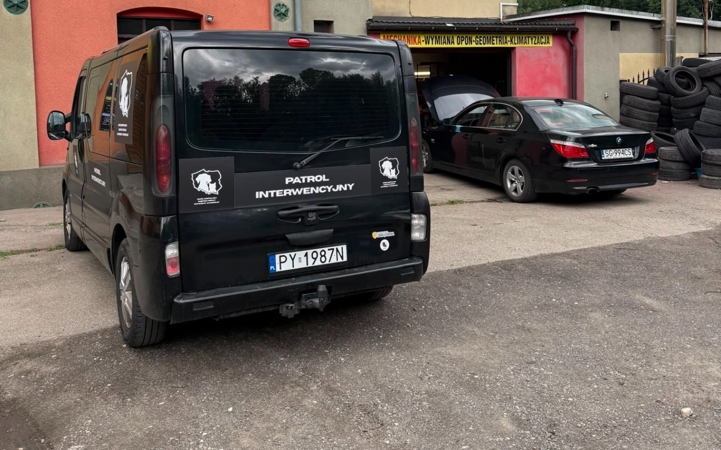 
        Czarny samochód zaparkowany na warsztatowej posesji obok v-wanu.
    