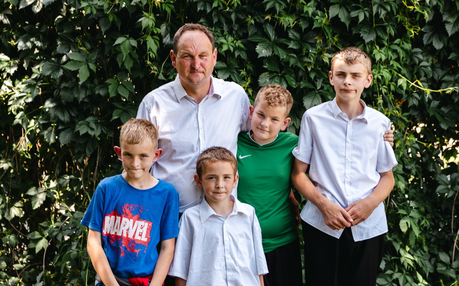 
        Grupa chłopców i mężczyzna na tle zieleni, wszyscy uśmiechnięci.
    
