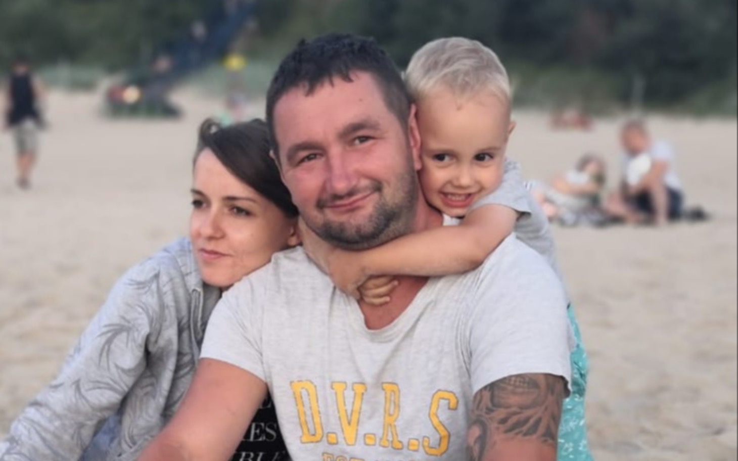 
        Rodzina na plaży: dorośli z dzieckiem na plecach, uśmiech i radość.
    