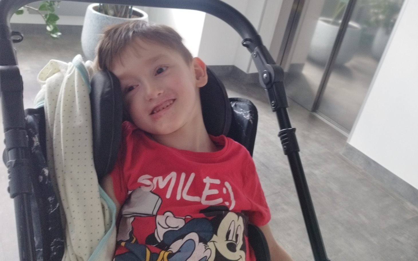 
        Dziecko na wózku, uśmiech na twarzy, w kolorowym ubraniu z postacią Mickey Mouse.
    