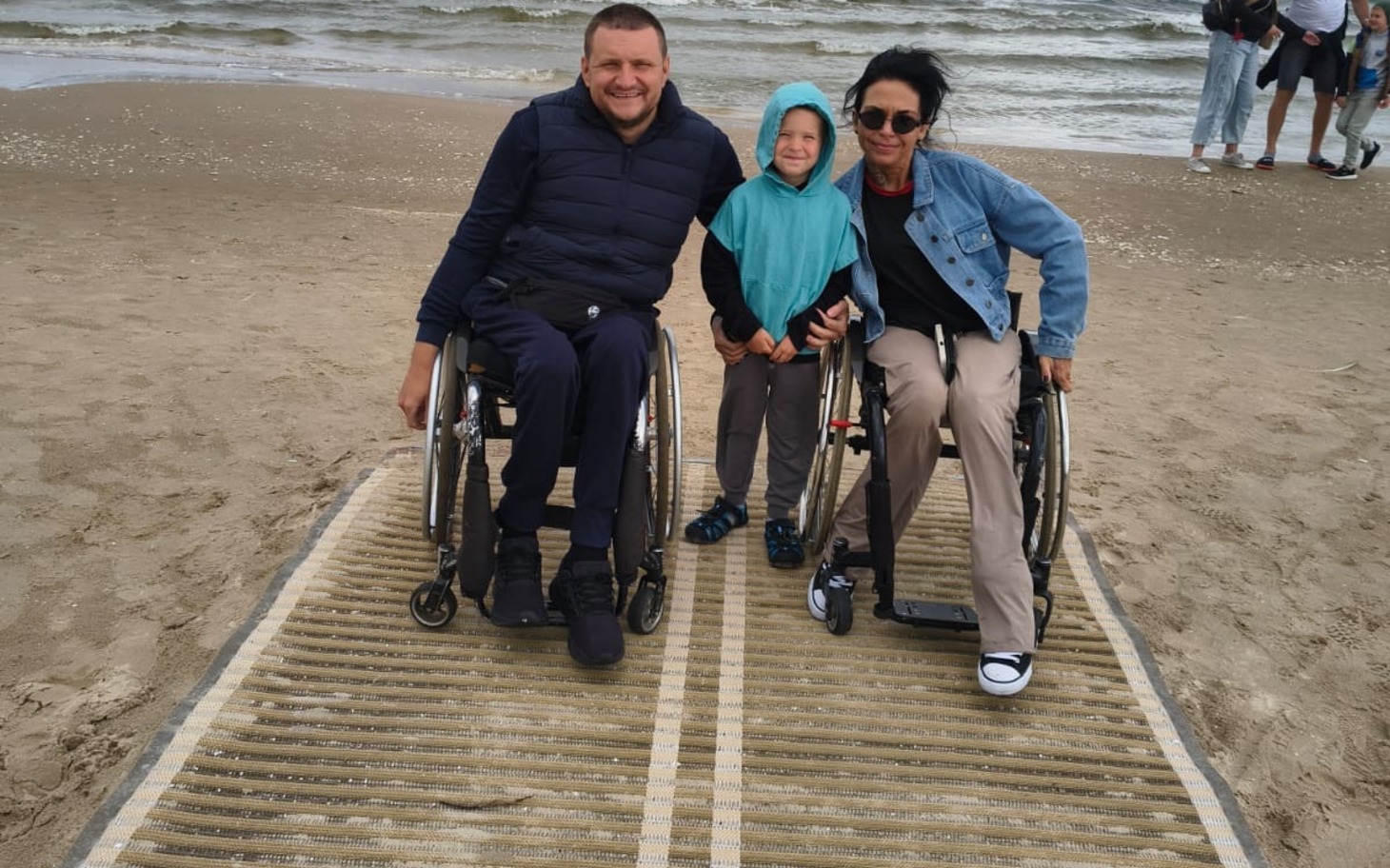 
        Troje osób na plaży, na wózkach inwalidzkich, trzymają się razem i uśmiechają
    