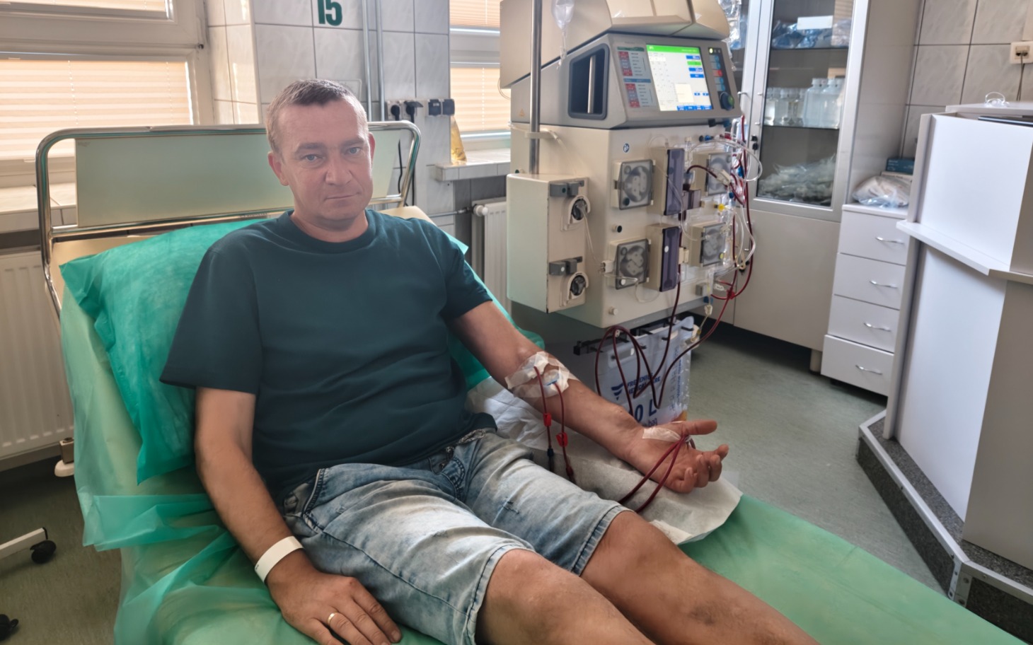 
        Chory mężczyzna w szpitalnym łóżku, na sali z aparaturą dializacyjną w tle.
    