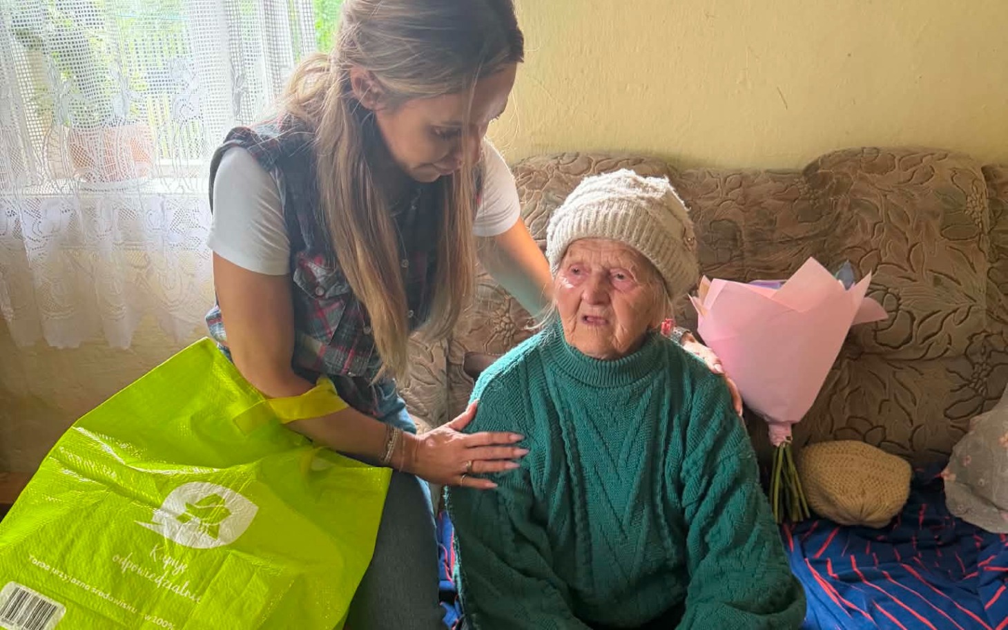 
        Opiekunka rozmawia z seniorką w domu, w tle bukiet róż
    