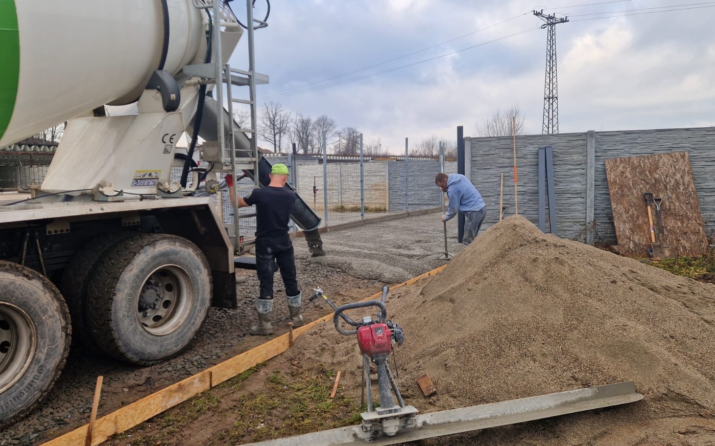 
        Robotnicy pracują przy gruncie, wóz betonowy i koparka w tle.
    