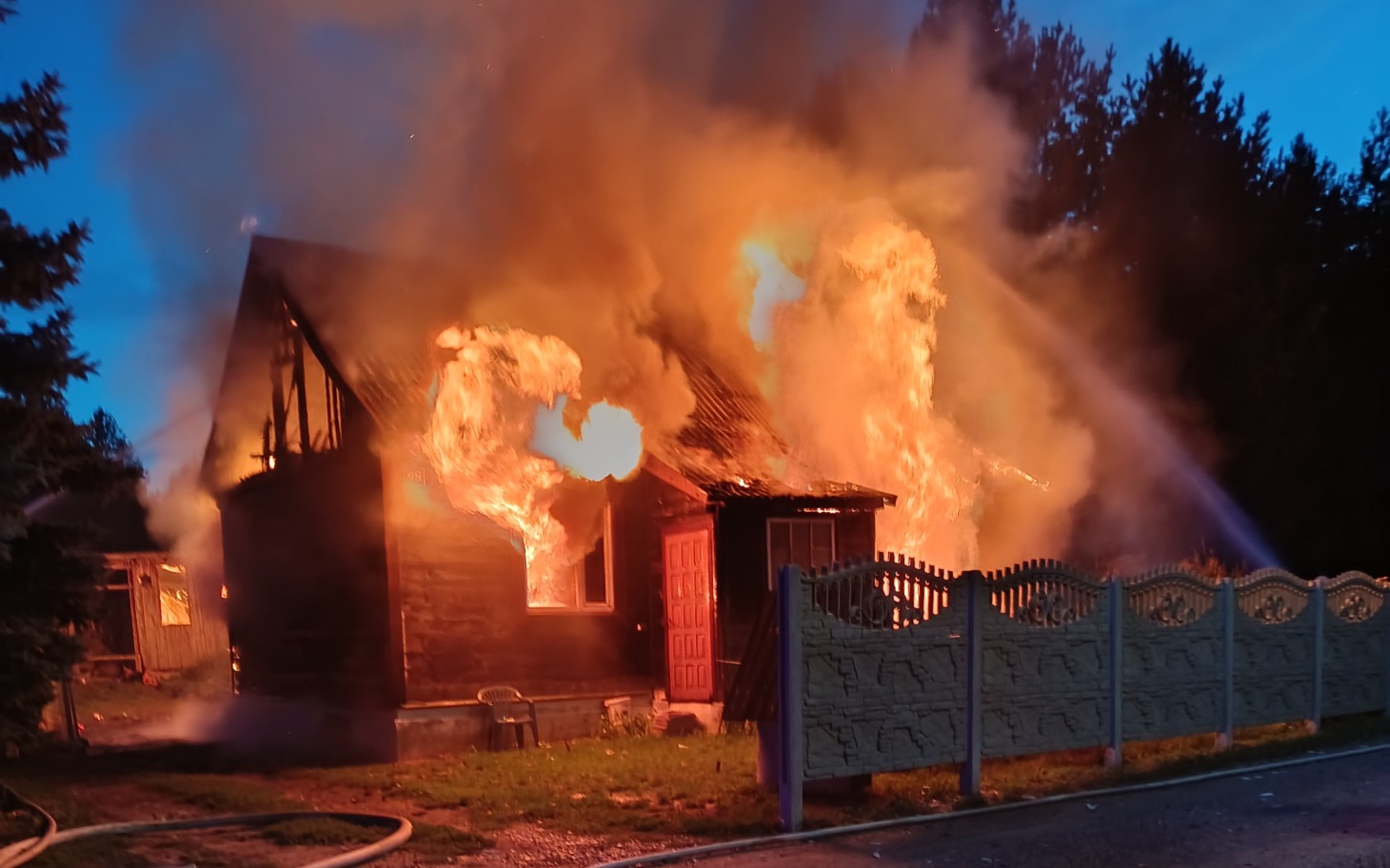 
        Płomienie pożaru nad domem, widoczny ogień i dym wokół drewnianej konstrukcji
    