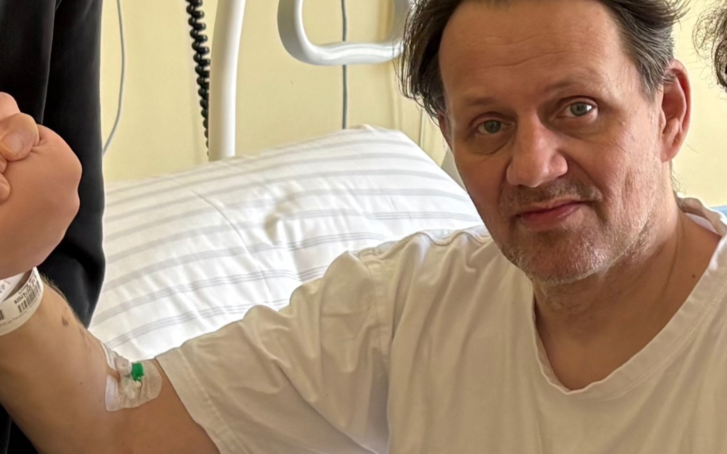 
        Mężczyzna na oddziale szpitalnym uśmiecha się do kamery, z opaską na nadgarstku.
    