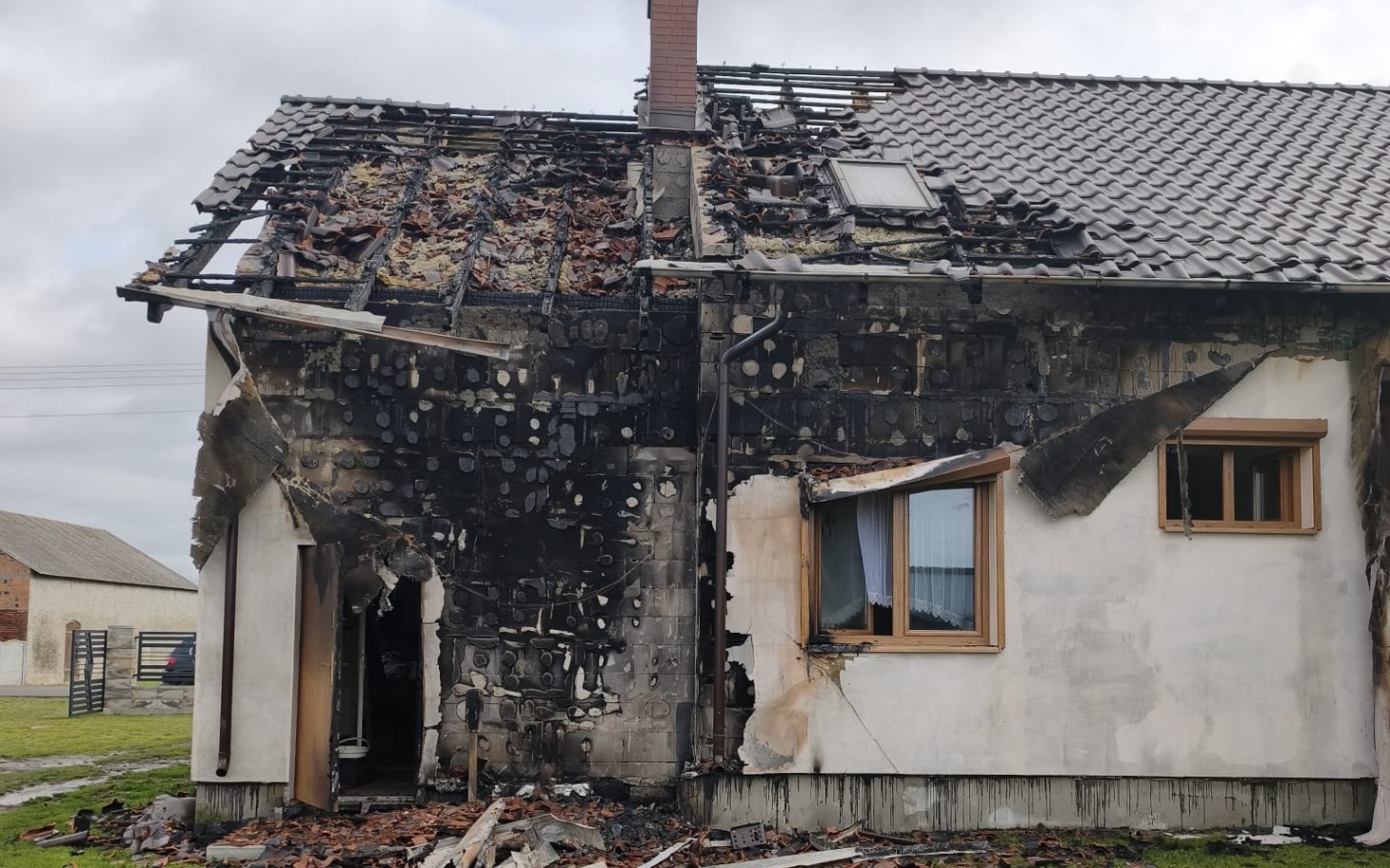 
        Zniszczony dom po pożarze z widoczną czarną ścianą i połamaną konstrukcją.
    