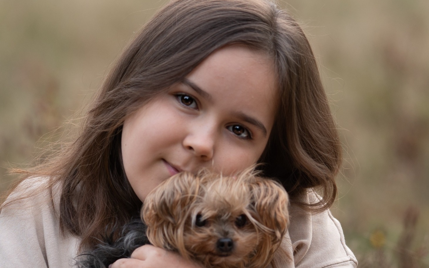 
        Dziewczynka trzymająca małego psa, uśmiechnięta na tle natury.
    